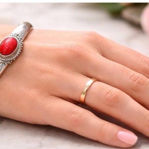 Può includere: Un bracciale rigido in argento lucido con una pietra rossa ovale centrale incastonata in una cornice decorativa in argento perlato, abbinato a un semplice anello a fascia liscio color oro. Il bracciale ha una finitura metallica riflettente.