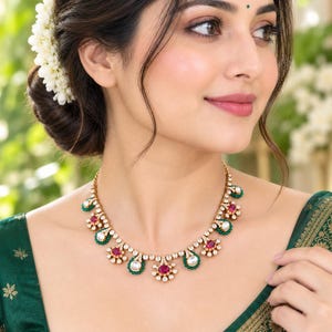 May include: A gold-toned necklace featuring a series of alternating oval-shaped red and white stones, each surrounded by small green beads and clear crystals. The piece is designed with a repeating floral-inspired pattern along the chain.