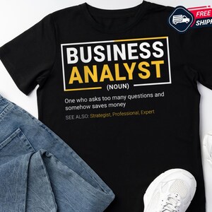 Peut inclure: T-shirt noir avec l'inscription "BUSINESS ANALYST" dans un design blanc et jaune. Le t-shirt comprend une définition et des termes connexes. Un jean bleu et des baskets blanches sont également dans l'image.