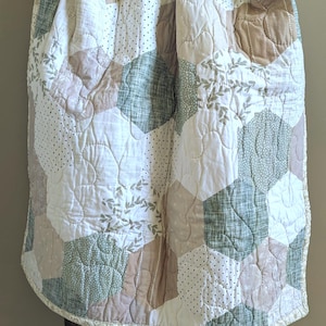 May include: A handmade quilt with a patchwork design. The quilt features a variety of hexagonal shapes in white, beige, and green. Some hexagons have dotted patterns or leaf motifs. The quilt is draped over a dark brown wooden frame.