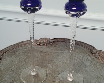 May include: Two elegant glass candle holders. Each features a deep blue bowl-shaped top, a decorative midsection, and a clear, textured stem. The candle holders are displayed on a decorative, silver-toned tray.