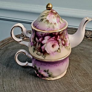 May include: A stacked teacup and teapot set. The teapot and teacup are decorated with pink roses and green leaves on a cream and purple background. The teapot has a gold finial and trim. The teacup has a handle and gold trim.