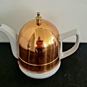 May include: A copper-colored teapot with a white handle, spout, and base. The lid has a small brass knob. The copper surface reflects the surroundings. The teapot is on a black surface.