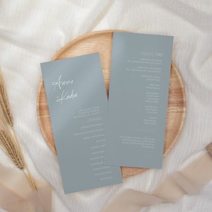 Könnte beinhalten: Zwei hellblaue Hochzeitseinladungen mit weißer Kalligrafie. Die Einladung links lautet "Aurora & Koden" und die Einladung rechts lautet "Wedding Party". Die Einladungen liegen auf einem Holzteller mit einem beigefarbenen Band und Weizenähren.