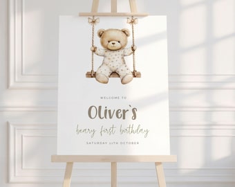 May include: A welcome sign for a first birthday celebration. The sign features a watercolor illustration of a teddy bear on a swing. The text reads "Welcome to Oliver's beary first birthday" and the date.