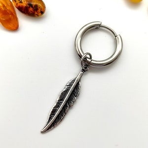 Può includere: Orecchino a cerchio color argento con un ciondolo a forma di piuma. La piuma presenta venature dettagliate e l'orecchino è posto su uno sfondo bianco con pietre color ambra.