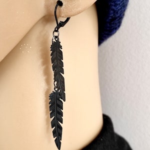 Könnte beinhalten: Schwarze Federn-Ohrringe mit langem, herabhängendem Design. Die Ohrringe sind aus Metall und haben eine matte Oberfläche.