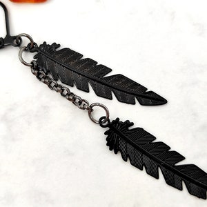 May include: Black feather earrings with a chain connecting the two feathers. The earrings are made of metal and have a black finish.