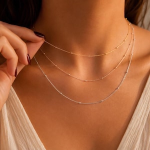 May include: A layered necklace set consisting of three delicate chains. Two chains are gold-toned and one chain is silver-toned. Each thin chain features small, evenly spaced metallic beads along the length of the strand, creating a simple, minimalist look.