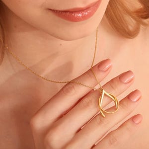 Op de afbeelding: Een goudkleurige ketting met een druppelvormige hanger. De hanger heeft een lusvormig ontwerp. De ketting wordt door een persoon vastgehouden. De achtergrond is een zachte, neutrale kleur.