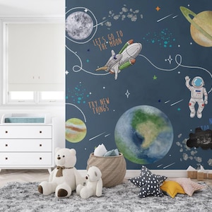 以下が含まれることがあります： 宇宙をテーマにした、青色の壁紙が特徴的な子供部屋。惑星、ロケット、宇宙飛行士が描かれています。「Let's Go To The Moon」、「Try New Things」、「Never Stop Exploring」の文字も確認できます。白いチェスト、グレーのラグ、ぬいぐるみも配置されています。