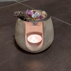 Könnte beinhalten: Ein grauer Beton-Ölbrenner mit silbernem Deckel und einer kleinen weißen Kerze im Inneren. Getrocknete Blumen werden oben in den Brenner gelegt.