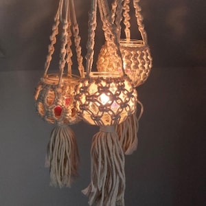 Könnte beinhalten: Drei Makramee-Kerzenhalter mit Glasbehältern und weißen Quasten. Die Kerzenhalter hängen von der Decke und die Kerzen sind angezündet.