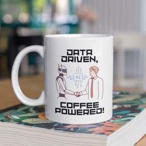 May include: White ceramic mug with a black design featuring a robot and a person shaking hands. The text on the mug reads "Data Driven, Coffee Powered!"