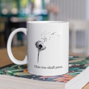 May include: White ceramic mug with a black and white dandelion illustration and the words "This too shall pass". The mug has a curved handle. The mug is sitting on a book with a patterned cover.