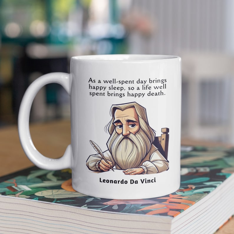 May include: White ceramic mug with a black and white illustration of Leonardo da Vinci. The mug features a quote by da Vinci: "As a well-spent day brings happy sleep, so a life well spent brings happy death."