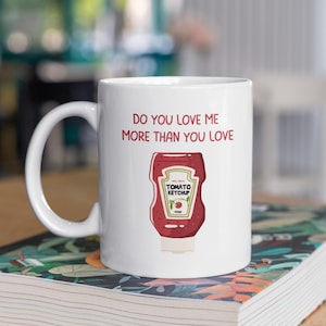 May include: White ceramic mug with a red and white illustration of a ketchup bottle and the text "Do you love me more than you love ketchup?"