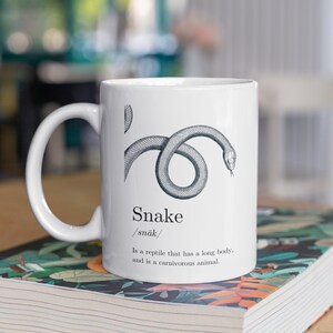 Könnte beinhalten: Eine weiße Keramiktasse mit einer Schwarzweißillustration einer Schlange. Die Tasse trägt das Wort "Snake" und die phonetische Schreibweise "/snāk/" aufgedruckt. Der Text unter der Illustration lautet: "Is a reptile that has a long body, and is a carnivorous animal."