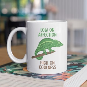 Puede incluir: Taza de cerámica blanca con una ilustración de camaleón verde y el texto "Low on Affection High on Coolness".