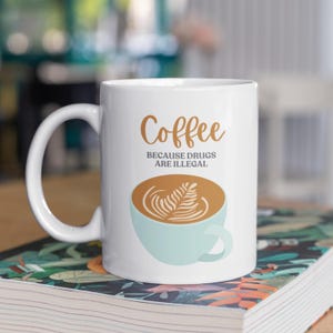 Peut inclure: Une tasse à café en céramique blanche avec un motif de café brun et blanc et le texte "Coffee because drugs are illegal."