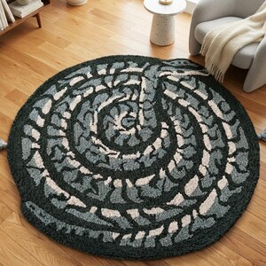 Può includere: Un tappeto rotondo trapuntato con un motivo a spirale nei colori grigio antracite, blu ardesia e crema. Il tappeto presenta una superficie testurizzata a pelo alto ed è rifinito con due nappe grigie abbinate sui lati opposti del bordo.