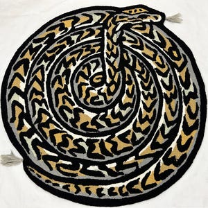 May include: A circular tufted rug featuring a coiled snake design in black, grey, and tan. The snake is depicted in a spiral pattern with a textured surface and two small grey tassels attached to the outer edge of the circular rug border.