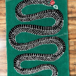 May include: A rectangular green rug featuring a large, winding black and white snake with a textured scale pattern. The snake is depicted with its mouth open, holding a small red and white object. The text Handloom Decor appears on the rug.
