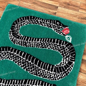 Puede incluir: Una alfombra rectangular verde que presenta una gran serpiente con patrón en blanco y negro enroscada en forma de letra S. La serpiente tiene una lengua roja que sobresale de su boca. El texto Handloom Decor es visible en las esquinas.