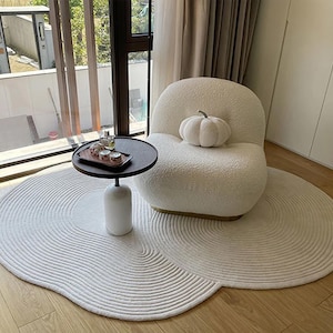 Puede incluir: Un sillón blanco y esponjoso con una calabaza blanca falsa en el asiento. El sillón está sobre una alfombra blanca y esponjosa con un patrón circular. Una pequeña mesa de madera con una bandeja de vidrio y tres botellas pequeñas está delante del sillón.