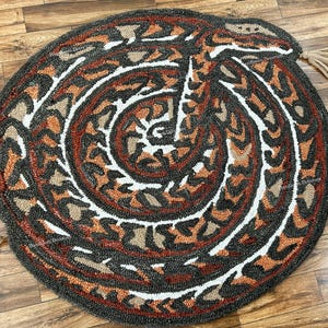May include: A circular rug featuring a coiled snake design in shades of dark grey, burnt orange, and cream. The textured surface displays a repeating geometric pattern throughout the body of the snake, finished with two beige tassels on the edges.