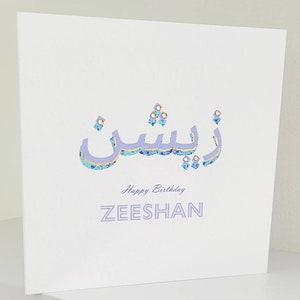 May include: A white card with a purple and gold design that says "Happy Birthday ZEESHAN" in a decorative font. The design includes Arabic calligraphy in purple and gold.