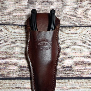 May include: A brown leather sheath with a black horseshoe tool inside. The sheath has a Maker brand logo embossed on the front.
