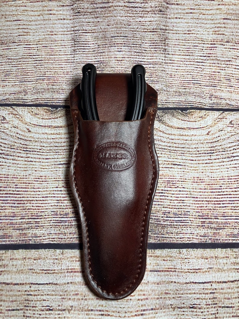 May include: A brown leather sheath with a black horseshoe tool inside. The sheath has a Maker brand logo embossed on the front.