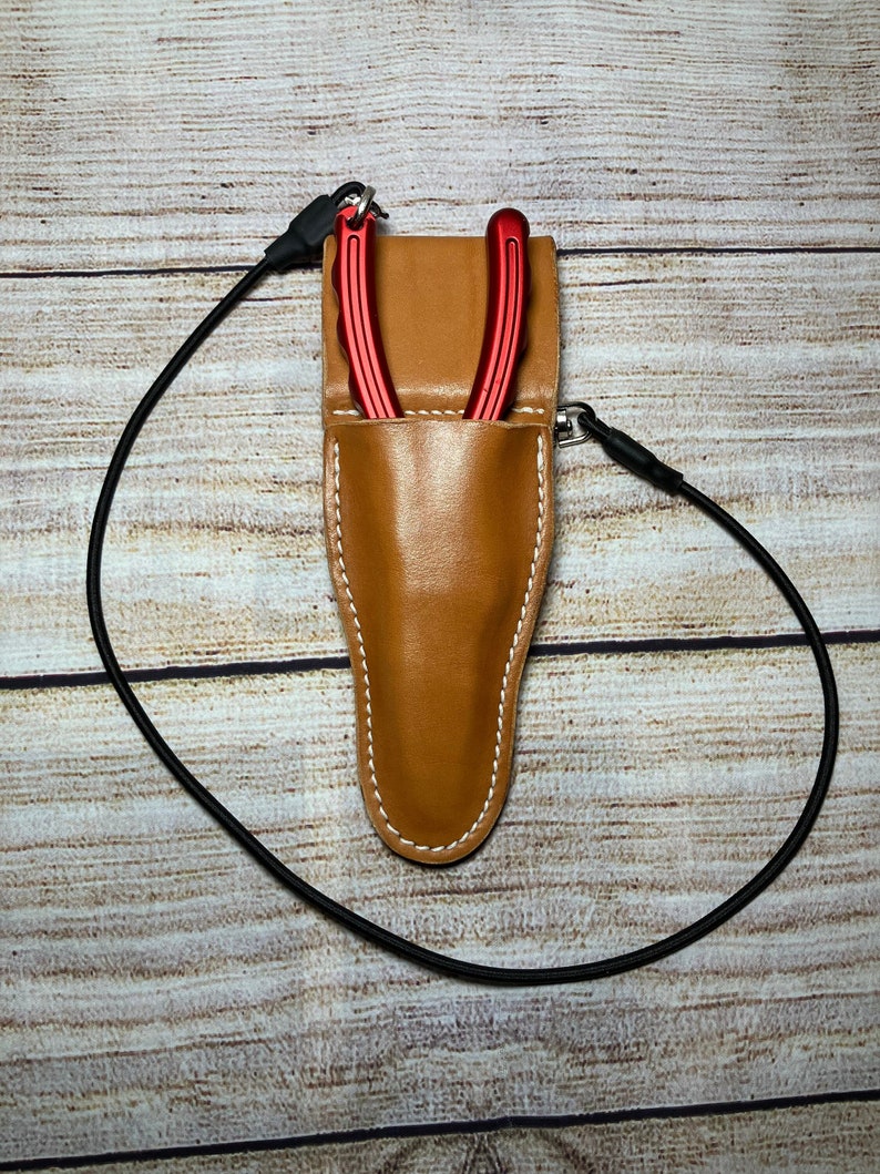 May include: A brown leather sheath with a red tool inside. The sheath has a white stitch detail and a black strap.