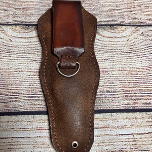 May include: A brown leather sheath with a belt loop and a metal ring. The sheath is designed to hold a small knife or tool.