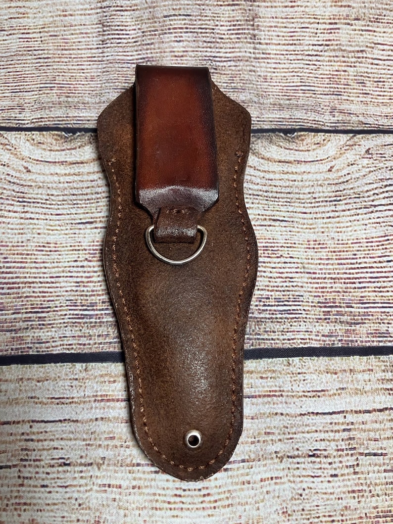 May include: A brown leather sheath with a belt loop and a metal ring. The sheath is designed to hold a small knife or tool.