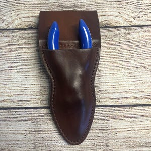 May include: A dark brown leather sheath with visible stitching along the edges, designed to hold a tool. Two bright blue, curved, smooth-finished handles of a pair of pliers or wire cutters are protruding from the top opening of the leather pouch.