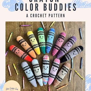 May include: A collection of twelve plush crochet crayon toys in various colors, each featuring a black stitched face and a black and white checkered band. The text CRAYON COLOR BUDDIES A CROCHET PATTERN is displayed above, with loose crayons scattered around the plushies.
