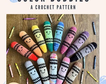 May include: A collection of twelve plush crochet crayon toys in various colors, each featuring a black stitched face and a black and white checkered band. The text CRAYON COLOR BUDDIES A CROCHET PATTERN is displayed above, with loose crayons scattered around the plushies.