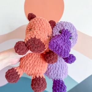 May include: Two plush stuffed animals made of chunky yarn. One is peach-colored with dark brown accents on the feet, ears, and snout. The other is purple with a darker purple bill and feet. Both have small black eyes and a soft, textured surface.