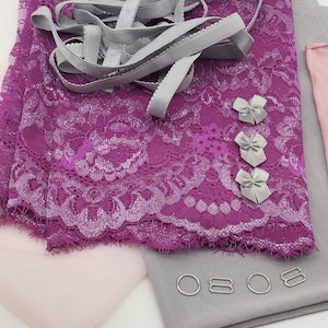 Op de afbeelding: Een verzameling benodigdheden voor het maken van lingerie. De afbeelding toont een levendige paarse kanten stof, grijs elastiek, kleine zilveren strikjes en metalen fournituren. Er zijn ook roze en grijze stoffen aanwezig, wat wijst op een doe-het-zelf lingeriemakerij.