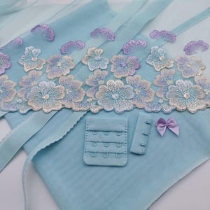 Könnte beinhalten: Hellblaues Dessous-Set mit floraler Stickerei in Weiß, Blau und Lila. Das Set enthält einen BH, Haken- und Ösenverschlüsse und eine kleine Lavendelschleife. Der Stoff ist zart und transparent.