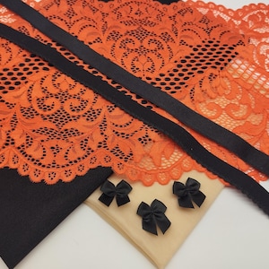 May include: An assortment of lingerie components, including orange lace fabric, black satin ribbon, and small black satin bows. The lace features a detailed floral pattern. The items are arranged on a neutral background, showcasing the textures and colors.