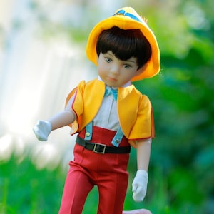 May include: A porcelain doll dressed as Peter Pan, wearing a yellow hat, a yellow jacket, red pants, and white gloves. The doll has brown hair and blue eyes.