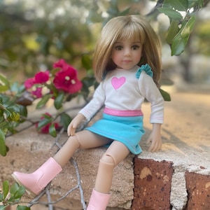 Puede incluir: Muñeca rubia de ojos marrones, vestida con una camiseta blanca con un corazón rosa, una falda azul y botas rosas. La muñeca está sentada en un muro de ladrillos, con flores rosas en el fondo.