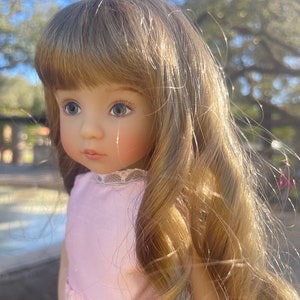 Puede incluir: Una muñeca con el pelo largo, ondulado y castaño claro, que lleva un vestido rosa con bordados florales blancos. La muñeca tiene ojos azules y una tez clara.