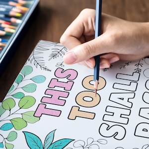 Puede incluir: Una p&aacute;gina para colorear con la frase "This Too Shall Pass" escrita en letras grandes y en negrita. La p&aacute;gina est&aacute; decorada con un patr&oacute;n de hojas y una mano sostiene un l&aacute;piz azul sobre la palabra "too".