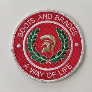 May include: Circular embroidered patch with a red backdrop and white edging. The words "BOOTS AND BRACES" and "A WAY OF LIFE" are in white. A gold helmet is at the centre, surrounded by a green laurel wreath.