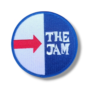 Puede incluir: Un parche redondo con un diseño dividido en blanco y azul. Una flecha roja apunta hacia la derecha. El texto "THE JAM" está en blanco, con una fuente estilizada en el lado azul. El parche tiene un borde azul.