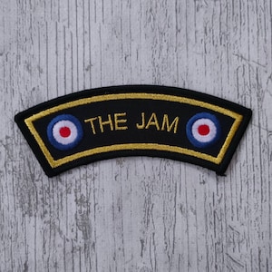 Puede incluir: Un parche negro arqueado con ribete dorado y el texto "THE JAM" en letras doradas. El parche presenta dos emblemas circulares con círculos concéntricos rojos, blancos y azules, que se asemejan a un objetivo.